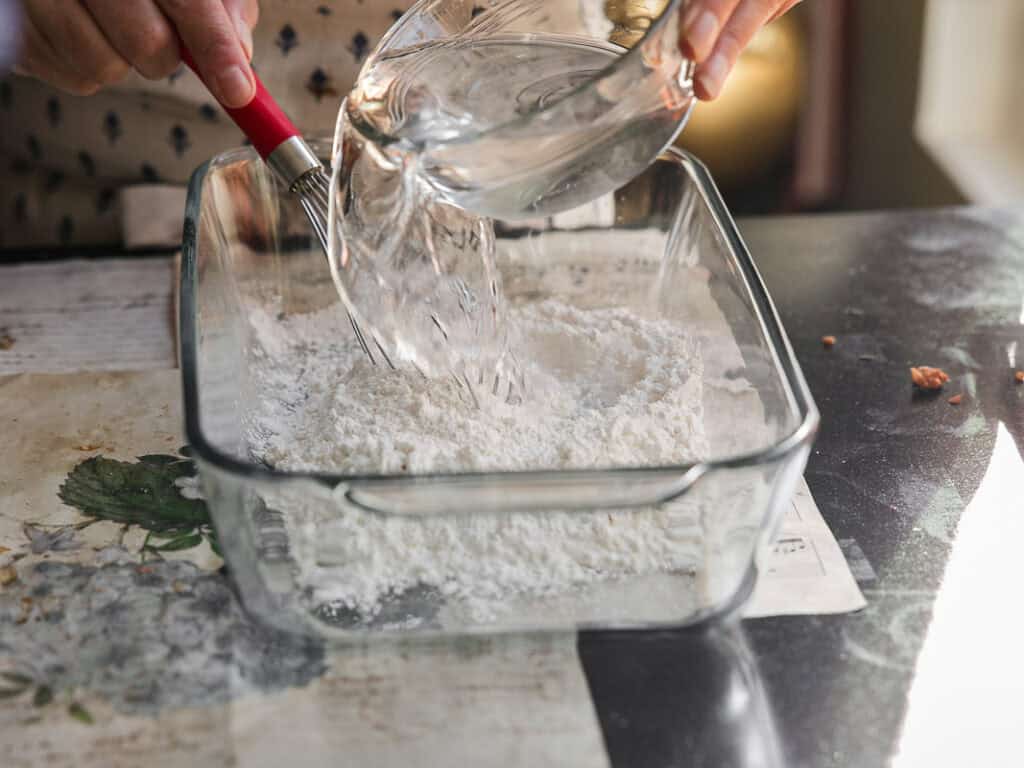 A person pours water from a glass bowl into a rectangular glass dish filled with flour. The scene is set on a table with a floral design.